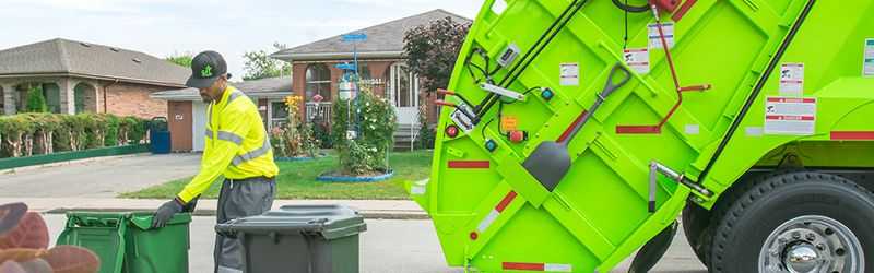 A man in a florescent shirt loading a green garbage bin into a florescent garbage truck